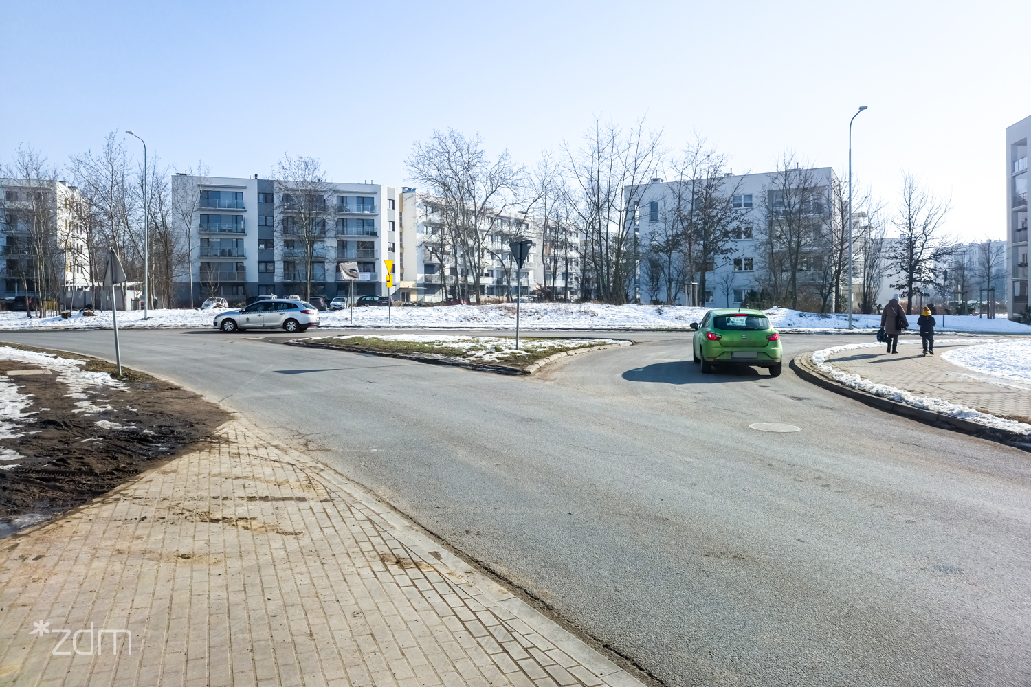 Zdjęcie skrzyżowania, które zmieni się w rondo, przejeżdżają dwa dwa samochody osobowe, w tle budynki