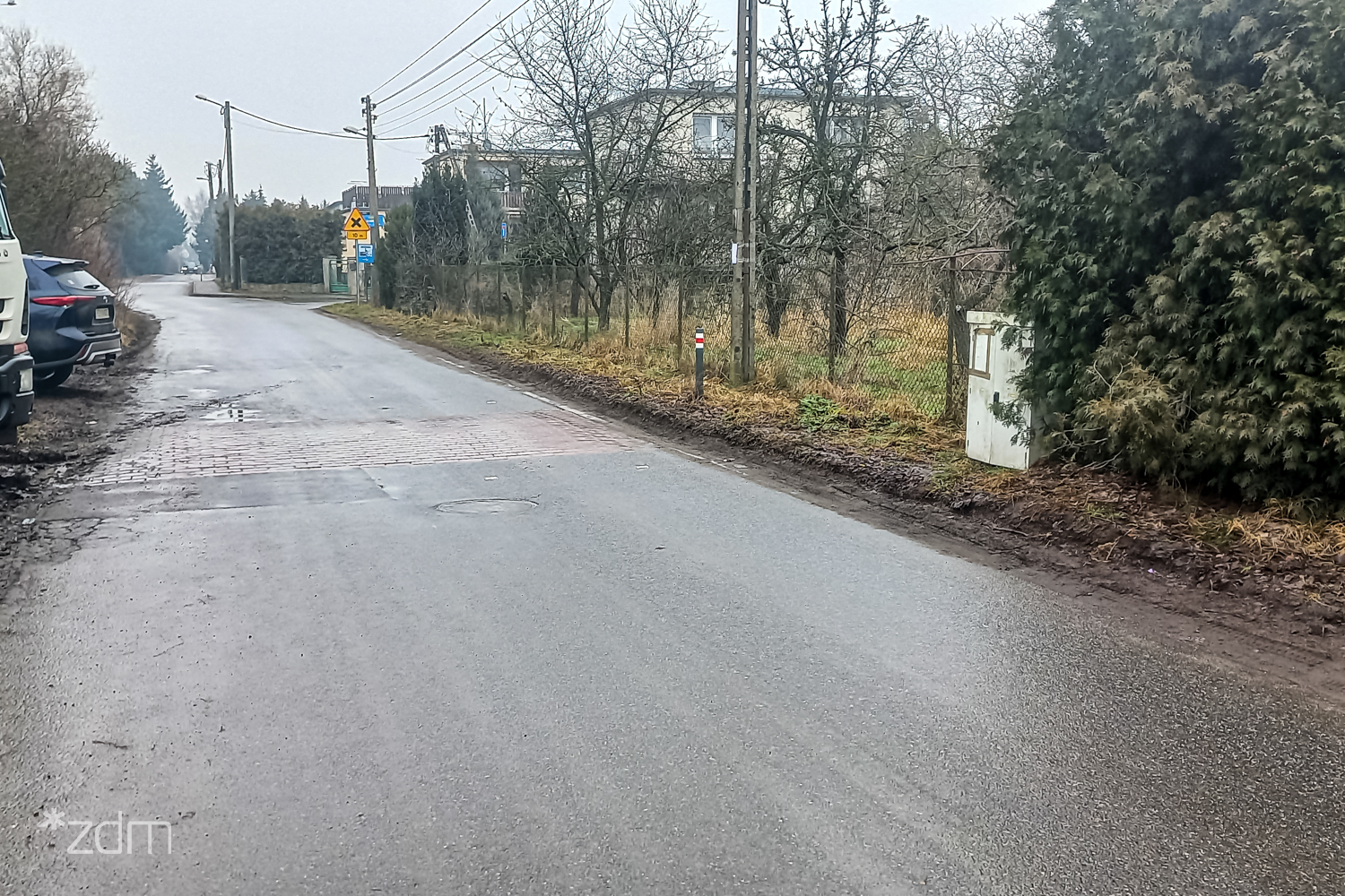 Zdjęcie ulic z Pokrzywna przed przebudową. Jezdnia asfaltowa bez chodnika, obok zieleń przyuliczna.