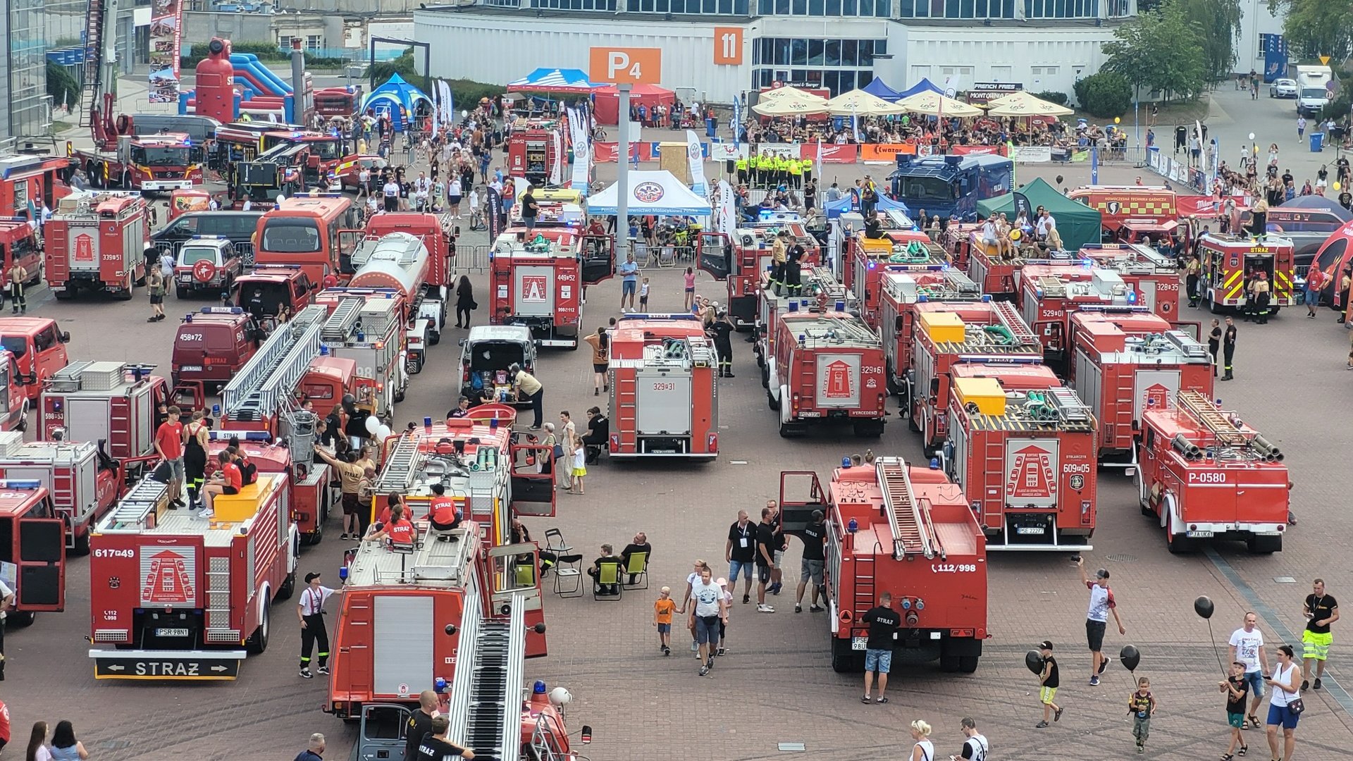 .Zdjęcie z góry na ustawione wozy strażackie na placu św. Marka na Międzynarodowych Targach Poznańskich. Wokół nich chodzą dorośli i dzieci. Niektórzy zaglądają do środka, inni robią sobie zdjęcia.