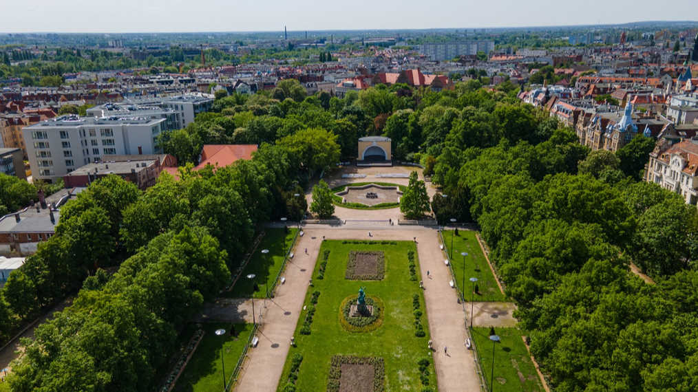 Zdjęcie z lotu ptaka przedstawiające Park Wilsona w Poznaniu, otoczony miejską zabudową. W centralnej części kadru znajduje się długa polana z trawnikami, alejkami spacerowymi oraz pomnikiem. Prowadzi ona w stronę placu z fontanną w kształcie gwiazdy oraz muszli koncertowej. Cały teren porastają gęste, wysokie, mocno zazielenione drzewa, a w tle widoczne są dachy kamienic i miejskie budynki oraz niebo.