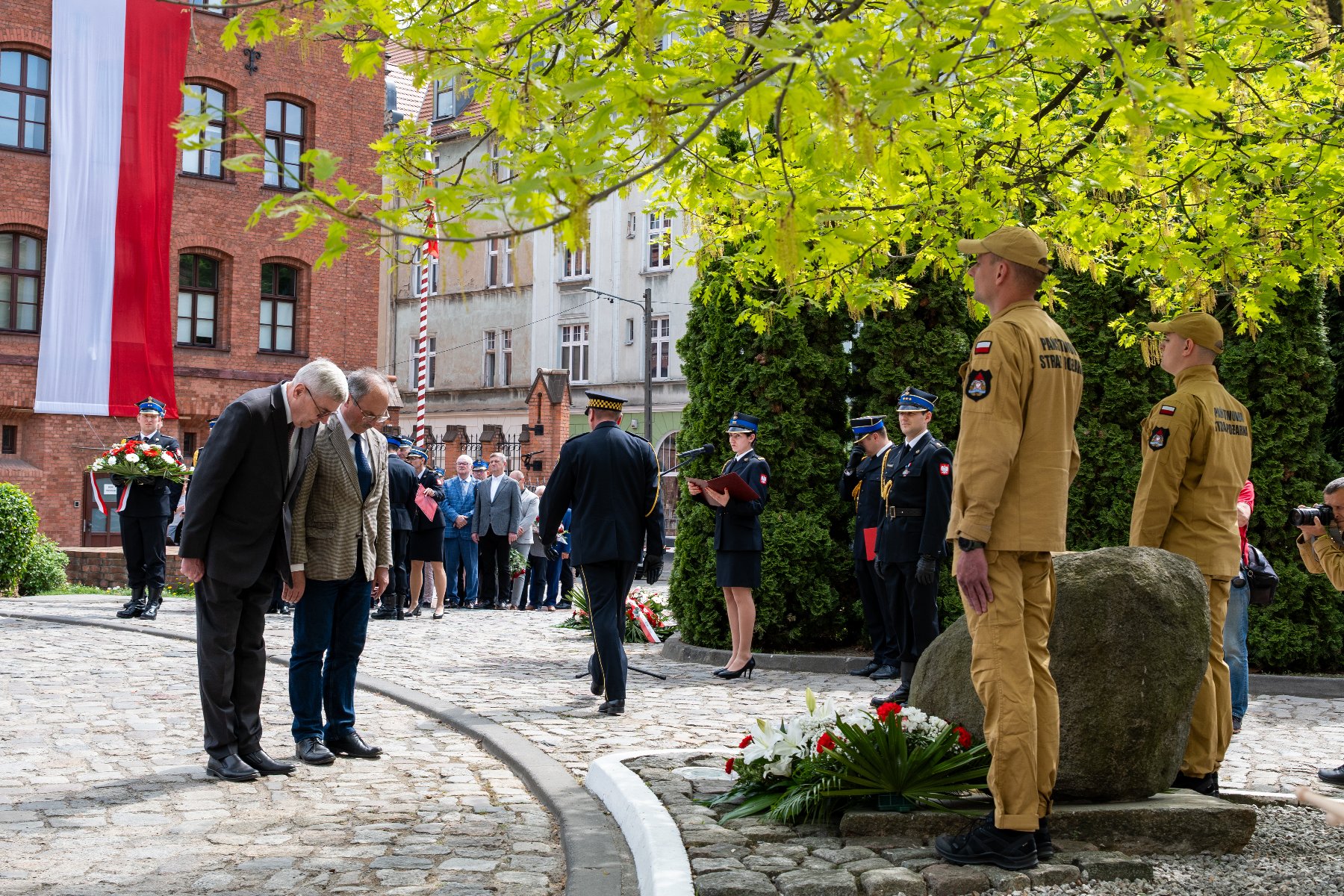 Zdjęcie z uroczystości, podczas której składane są kwiaty pod tablicą zamieszczoną na kamieniu. Sytuacja ma miejsce na zewnątrz, widać poczet sztandarowy, a także osoby składające kwiaty.