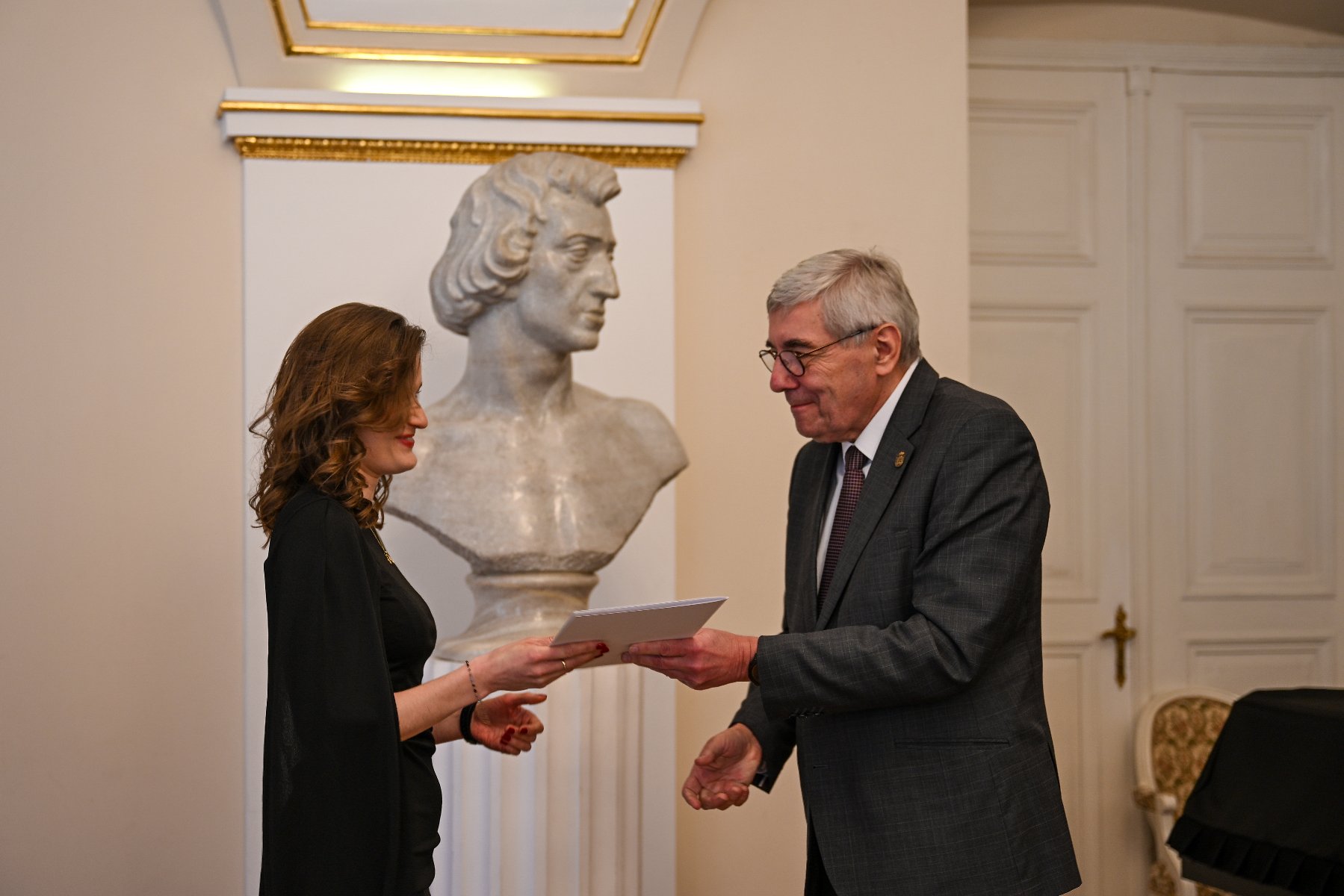 Zdjęcie z uroczystości wręczenia wyróżnień. Przewodniczący rady miasta gratuluje laureatom, wręczając im dyplomy. Sytuacja dzieje się w sali.