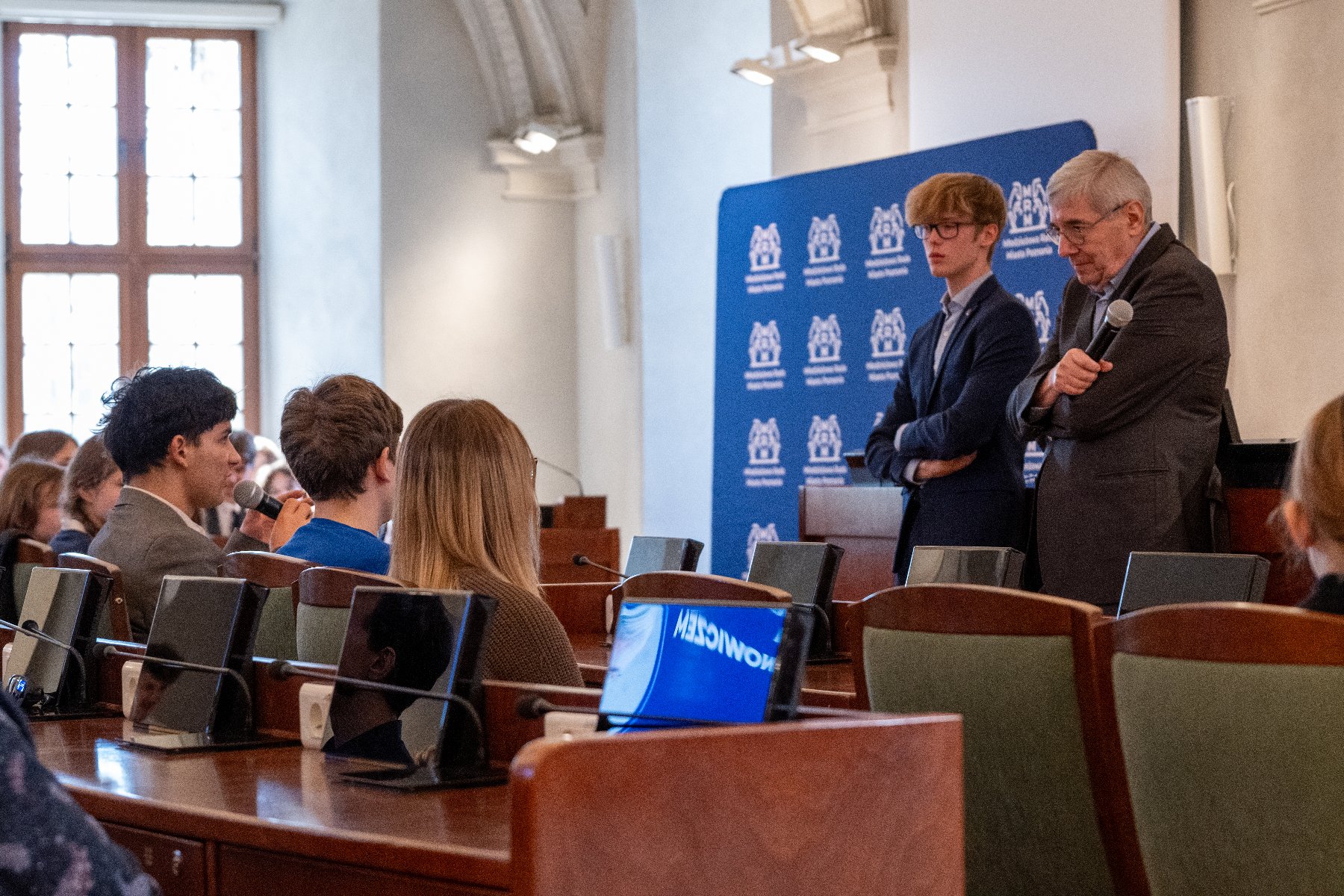 Zdjęcie z wydarzenia. W centralnym punkcie przewodniczący rady miasta, wokół siedzą młodzi ludzie.