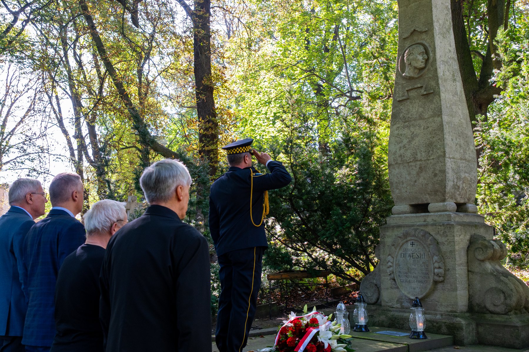 Zdjęcie ze cmentarza. Grupa mężczyzn w garniturach oddaje hołd nad kwiatami.
