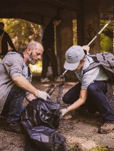 Dwie osoby kucają pod betonową konstrukcją wiaduktu, zbierając śmieci. Mężczyzna z lewej strony, w szarej bluzie i rękawiczkach, trzyma otwarty czarny worek. Młoda osoba obok niego, w jasnej czapce z daszkiem, jasnej koszulce i ciemnych spodniach, wkłada odpady do worka za pomocą chwytaka. W tle widać inne osoby idące ścieżką oraz zielone zarośla.