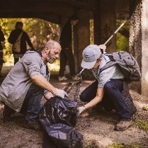 Dwie osoby kucają pod betonową konstrukcją wiaduktu, zbierając śmieci. Mężczyzna z lewej strony, w szarej bluzie i rękawiczkach, trzyma otwarty czarny worek. Młoda osoba obok niego, w jasnej czapce z daszkiem, jasnej koszulce i ciemnych spodniach, wkłada odpady do worka za pomocą chwytaka. W tle widać inne osoby idące ścieżką oraz zielone zarośla.