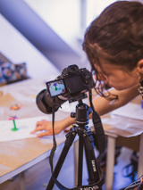 Młoda kobieta z ciemnymi, upiętymi włosami, pochylona nad profesjonalnym aparatem fotograficznym umieszczonym na czarnym statywie. Postać ubrana w czarną bluzkę patrzy na podgląd ekranu aparatu. Przed nią na stole widać fotografowany obiekt przypominający kwiata z plasteliny.