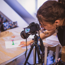 Młoda kobieta z ciemnymi, upiętymi włosami, pochylona nad profesjonalnym aparatem fotograficznym umieszczonym na czarnym statywie. Postać ubrana w czarną bluzkę patrzy na podgląd ekranu aparatu. Przed nią na stole widać fotografowany obiekt przypominający kwiata z plasteliny.