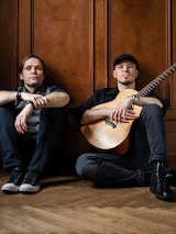 Dwóch mężczyzn siedzących na podłodze pod brązową, drewnianą ścianą z dekoracyjnymi kasetonami. Po lewej stronie mężczyzna z dłuższymi ciemnymi włosami, ubrany w szarą kamizelkę, koszulę i ciemne spodnie, z dłońmi opartymi na kolanach. Po prawej stronie mężczyzna w czapce z daszkiem, okularach i czarnym ubraniu, trzymający na kolanach jasną gitarę akustyczną. Obaj patrzą bezpośrednio w obiektyw z poważnym wyrazem twarzy.