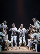 Scena ze spektaklu teatralnego przedstawiająca grupę aktorów w jasnych strojach na ciemnym tle. W centrum stoją dwaj mężczyźni w ekspresyjnych pozach, z uniesionymi dłońmi. Pozostali siedzą na drewnianych stołkach z białymi nóżkami.