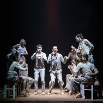 Scena ze spektaklu teatralnego przedstawiająca grupę aktorów w jasnych strojach na ciemnym tle. W centrum stoją dwaj mężczyźni w ekspresyjnych pozach, z uniesionymi dłońmi. Pozostali siedzą na drewnianych stołkach z białymi nóżkami.