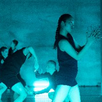 Kilka osób na scenie ubranych w czarne stroje. Scena jest przyciemniona i wypełniona niebieskim światłem.