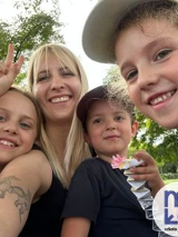 Czworo uśmiechniętych osób - troje dzieci i dorosła osoba - pozuje razem na tle zieleni w parku. Jedno z dzieci pokazuje znak pokoju, inne trzyma figurkę z papieru. W prawym dolnym rogu widoczne jest logo z niebieskim napisem "Miła" z czerwoną kropką nad "i", wpisaną w białe koło. Pod spodem napis "szkoła językowa".