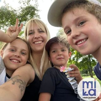 Czworo uśmiechniętych osób - troje dzieci i dorosła osoba - pozuje razem na tle zieleni w parku. Jedno z dzieci pokazuje znak pokoju, inne trzyma figurkę z papieru. W prawym dolnym rogu widoczne jest logo z niebieskim napisem "Miła" z czerwoną kropką nad "i", wpisaną w białe koło. Pod spodem napis "szkoła językowa".