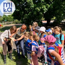 Grupa dzieci stoi na zewnątrz wokół dwóch dorosłych, którzy pokazują im mapę. Wszyscy mają plecaki, a w tle widać drzewa i słoneczną pogodę. W lewym górnym rogu widoczne jest logo z niebieskim napisem "Miła" z czerwoną kropką nad "i", wpisaną w białe koło. Pod spodem napis "szkoła językowa".
