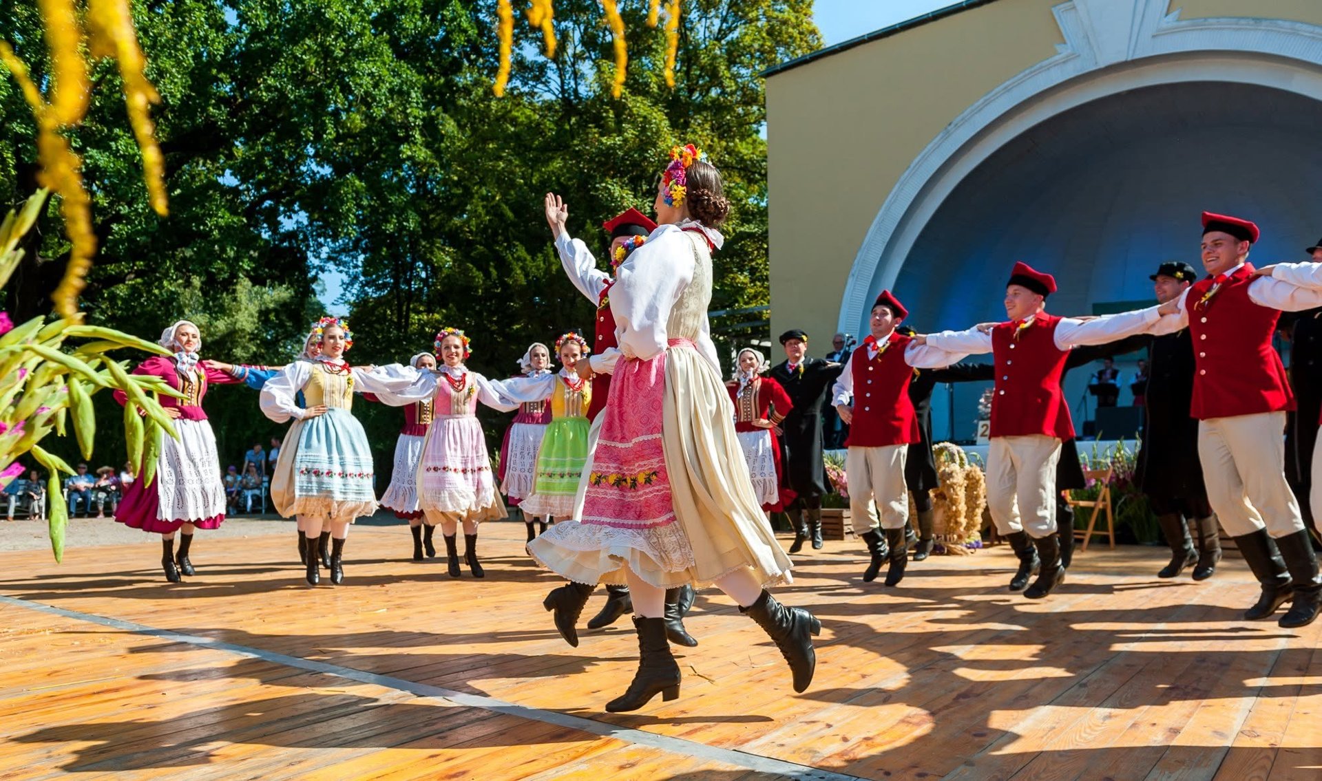 Zespół ludowy w tradycyjnych strojach wykonujący taniec na drewnianej scenie plenerowej podczas festiwalu.
