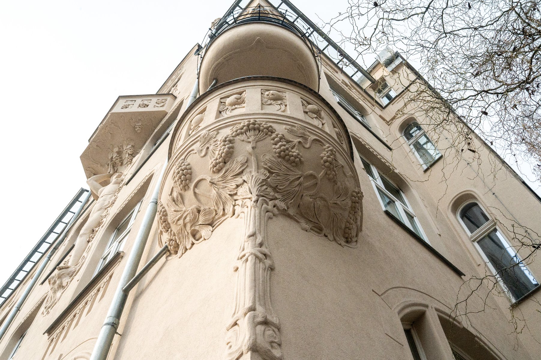 Zewnętrzna fasada budynku z bogato zdobionymi detalami architektonicznymi. Na zaokrąglonym narożniku znajduje się ornamentyka, w tym motywy roślinne przypominające winorośl. Budynek ma kilka okien, a fotografia została wykonana z perspektywy patrzenia w górę.