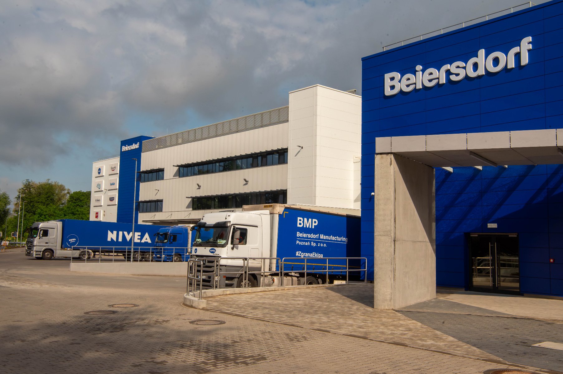 Zewnętrzny widok na budynki fabryki Beiersdorf w Poznaniu, z kilkoma ciężarówkami stojącymi na placu załadowczym. Na pierwszym planie niebieski budynek z logo "Beiersdorf".