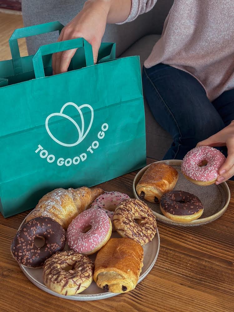 Zielona torba z napisem Too good to go, przed którą stoją dwa talerze wypełnione pączkami.
