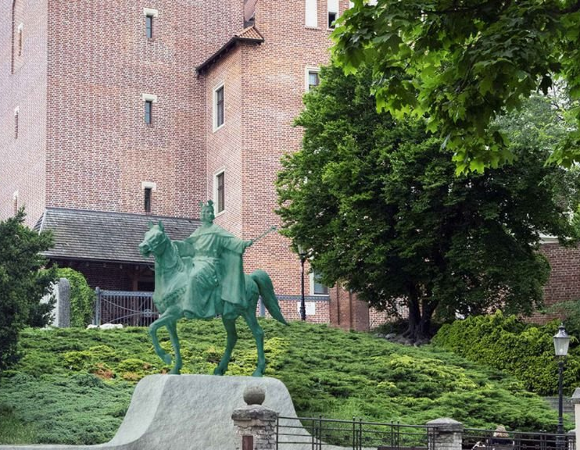 Zielony, metalowy pomnik na koniu stojący na szarym, kamiennym postumencie. Za pomnikiem wznosi się duża, ceglana ściana zabytkowego budynku z małymi oknami. Po prawej stronie widoczne drzewa i znak drogowy.