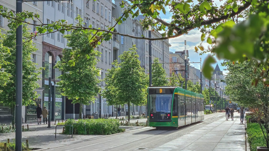 Zielony tramwaj numer 9 jedzie środkiem nowoczesnej miejskiej ulicy, otoczonej drzewami, zielenią i budynkami o zróżnicowanej architekturze. Na chodnikach widać przechodniów, a całość oświetla słońce.