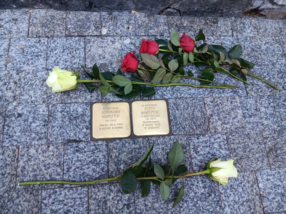 Złote kamienie z imionami i nazwiskami wmurowane w chodnik. Obok nich leżą białe i czerwone róże.