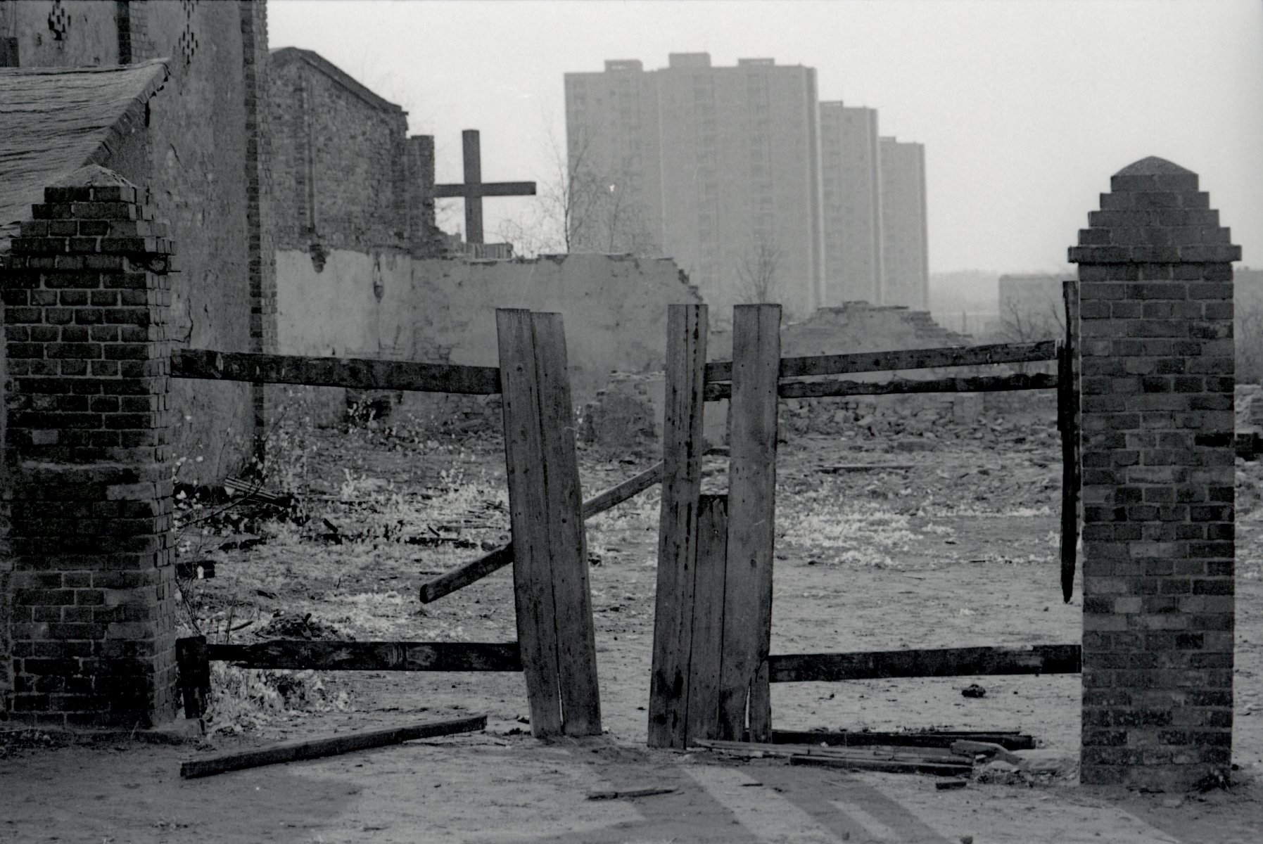 Zniszczona brama wjazdowa, podwórze zasypane gruzem, drewniany krzyż, w tle 16-piętrowe wieżowce