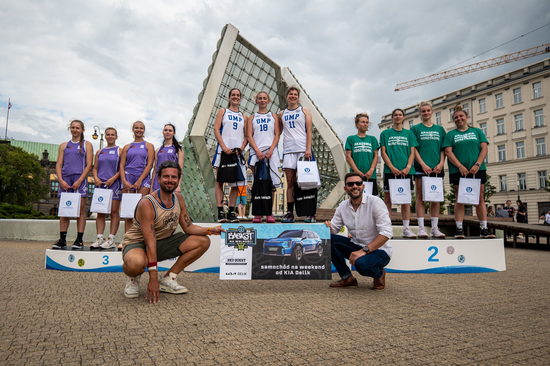Zwycięskie drużyny kategorii OPEN kobiet w imprezie Basket na Wolności, fot. Piotrek Przyborowski/Basket na Wolności - na tle fontanny stoją 3 drużyny, po bokach 4-osobowe, a w środku 3-osobowa. z białych strojach. Po lewej stronie mają fioletowe stroje, a po prawej zielone koszulki i czarne spodenki. Wszystkie trzymają w rękach nagrody rzeczowe. Z przodu kucają 2 mężczyźni trzymając baner