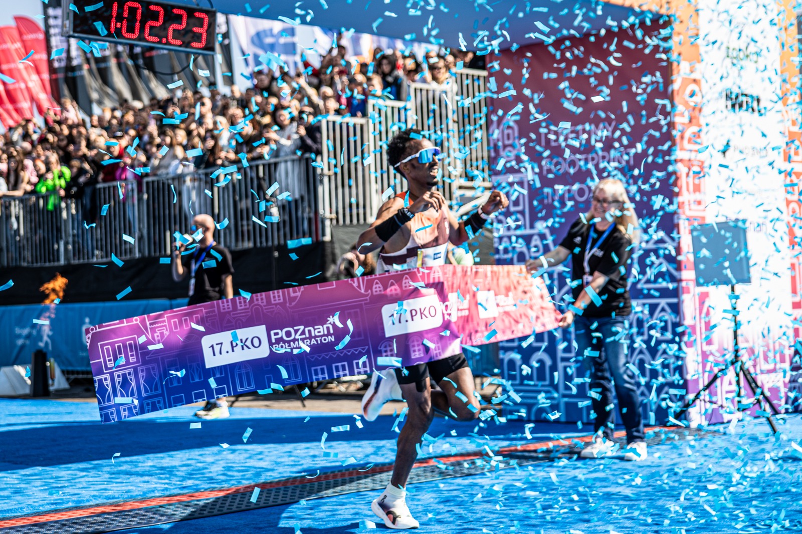 Zwycięzca 17.PKO Poznań Półmaratonu na mecie, fot. Klaudia Berda - mężczyzna w okularach przeciwsłonecznych wbiega na metę wśród konfetti. W tle widać trybunę z kibicami. Zawodnik wbiega po niebieskim dywanie.