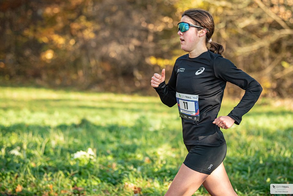 Zwyciężczyni pierwszego w nowym sezonie biegu City Trail w Poznaniu, fot. Piotr Oleszak - na tle trawy i lasu biegnie kobieta w czarnym stroju