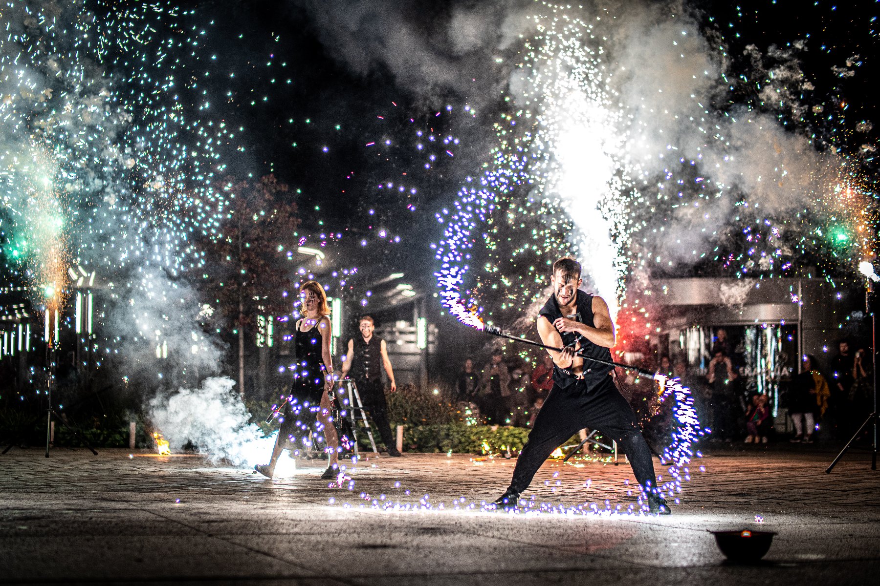Artyści wykonują pokaz fireshow nocą - kobieta i mężczyzna przeprowadzają pokaz z wykorzystaniem ognia, a wokół unoszą się iskry i fajerwerki, podczas gdy publiczność ogląda występ