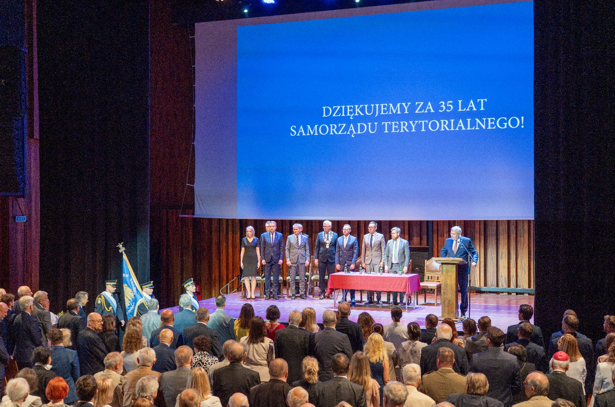 duża sala wypełniona ludźmi stojącymi pod secną, na scenie stoi około kilkunastu osób, jedna osoba stoi przy mównicy - grafika artykułu