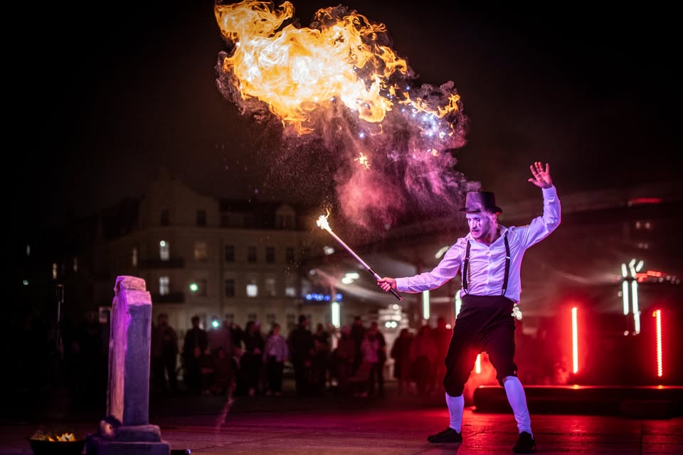 Element występu artystycznego. Mężczyzna ubrany w cylinder z pomalowaną na biało twarzą, trzyma w ręku pochodnię, a nad nim łuna ognia. Wydarzenie odbywa się nocą.