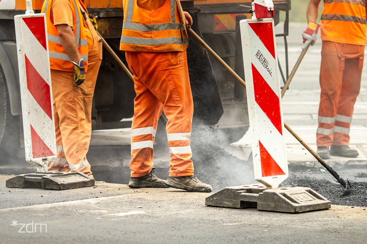 Fragment budowy drogi. Trzech pracowników ubranych w pomarańczowe spodnie i kamizelki odblaskowe stoi na asfalcie, trzymając szpadle lub grabie do wyrównywania nawierzchni. Na pierwszym planie dwa biało-czerwone słupki drogowe. Z lewej strony na dole widoczne małe, białe logo w cudzysłowie: "*zdm".