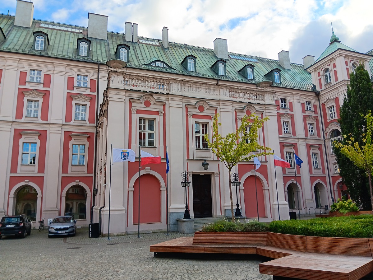 gmach od strony dziedzińca zabytkowa architektura przed wejście do gmachu ławki i zieleń oraz flagi na masztach flaga Poznania Polski i Unii Europejskiej