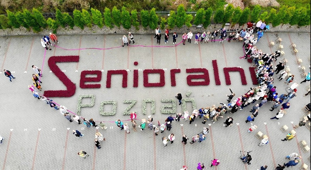 grafika przedstawia widok z góry na plac na którym ułożony został z kwiatów napis Senioralni