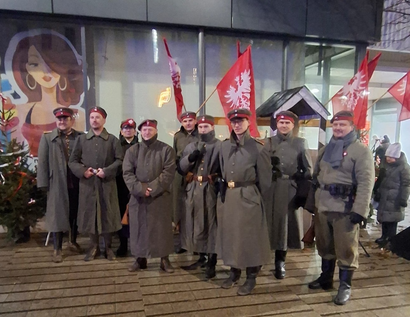 Grupa mężczyzn ubrana w mudury żołnierzy z epoki. W tole flagi Powstania Wielkopolskiego.