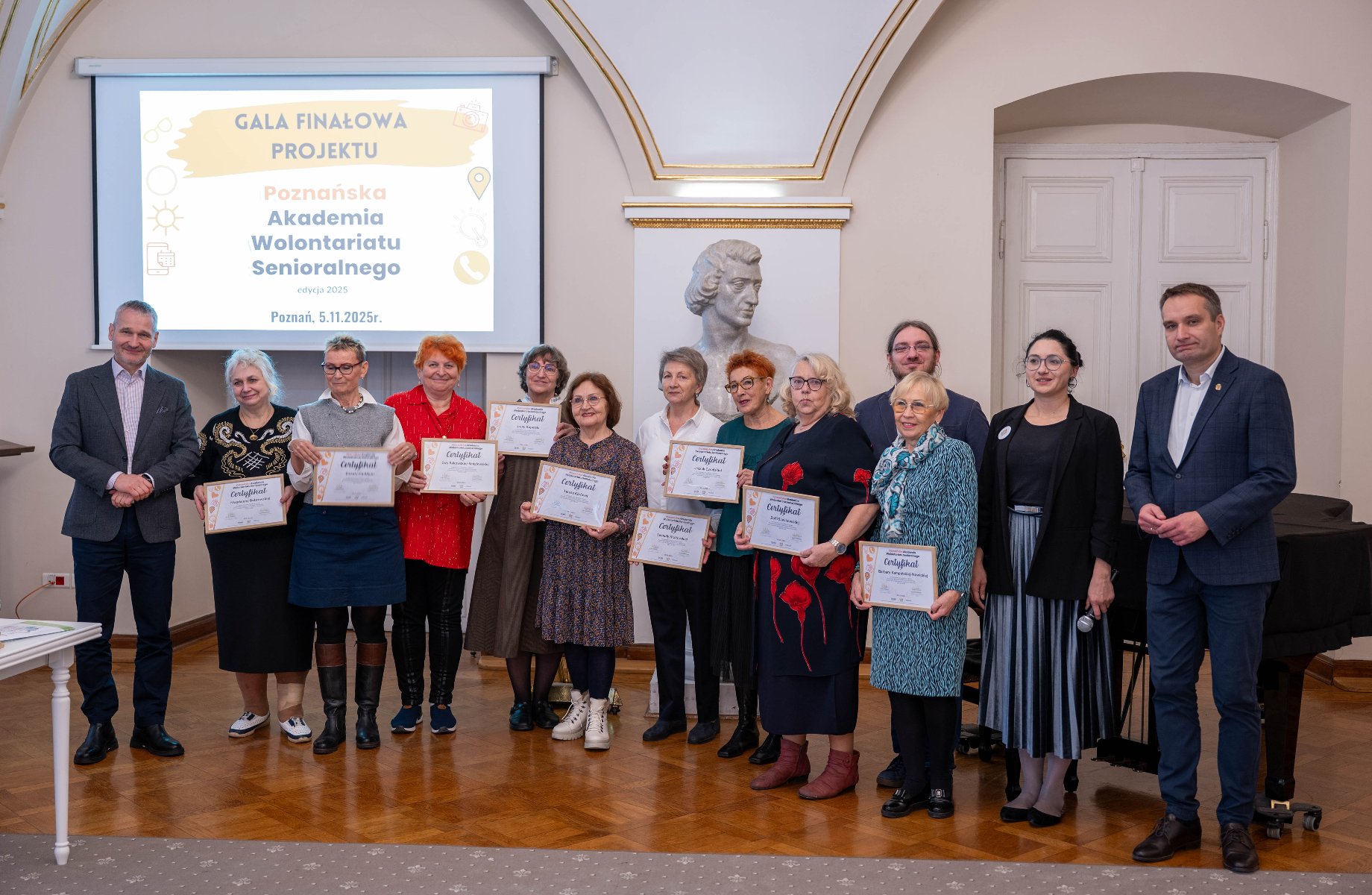 Grupa osób pozuje do zdjęcia w sali z drewnianą podłogą, podczas Gali Finałowej. W centrum ujęcia w rzędzie stoi dziesięć starszych kobiet, trzymających certyfikaty, z uśmiechem skierowanym w stronę aparatu.