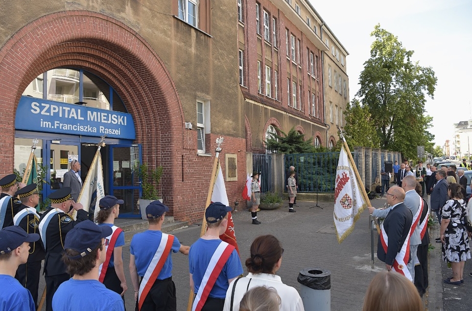grupa osób ze sztandarami i szarfami w biało czerwonych kolorach stoi przed wejściem do budynku szpitala