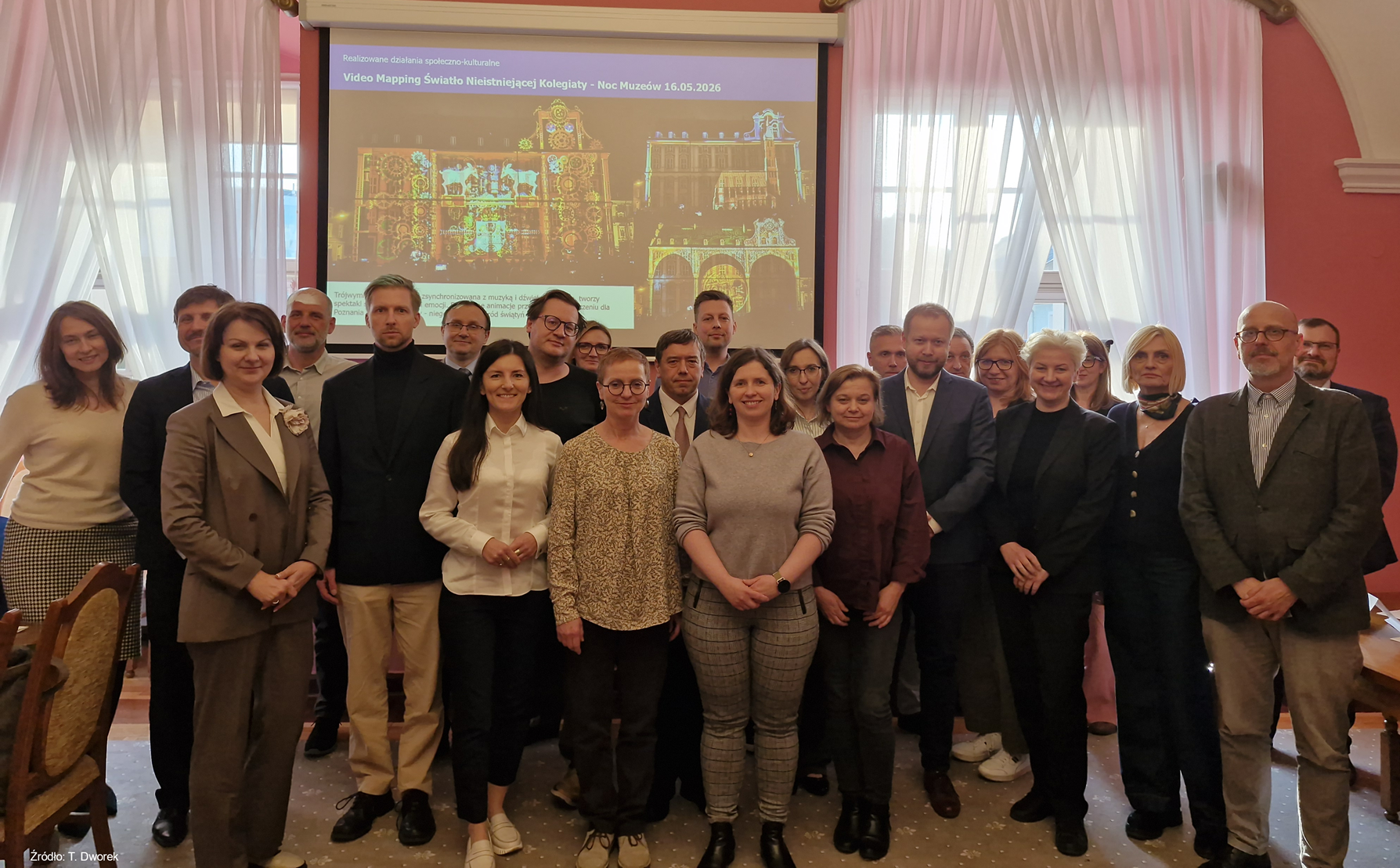 Kilkanaście osób stoi na tle ekranu, którym wyświetlana jest prezentacja multimedialna.