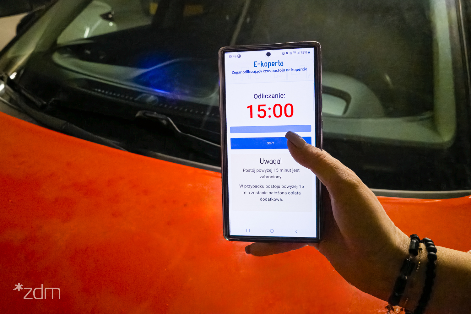 Kobieca ręka trzyma telefon z elektronicznym zegarem odmierzającym czas parkowania. W tle pomarańczowy samochód