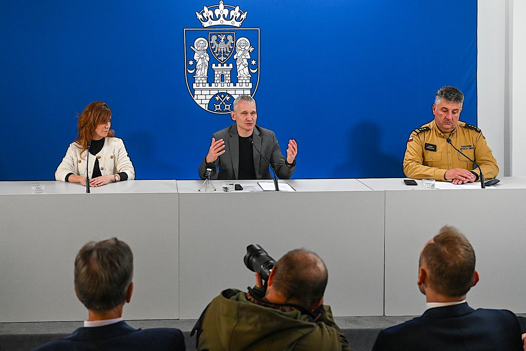 konferencja pradsowa - trzy osoby siedzą za stołem, w tle ścianka niebieska z herbem miasta