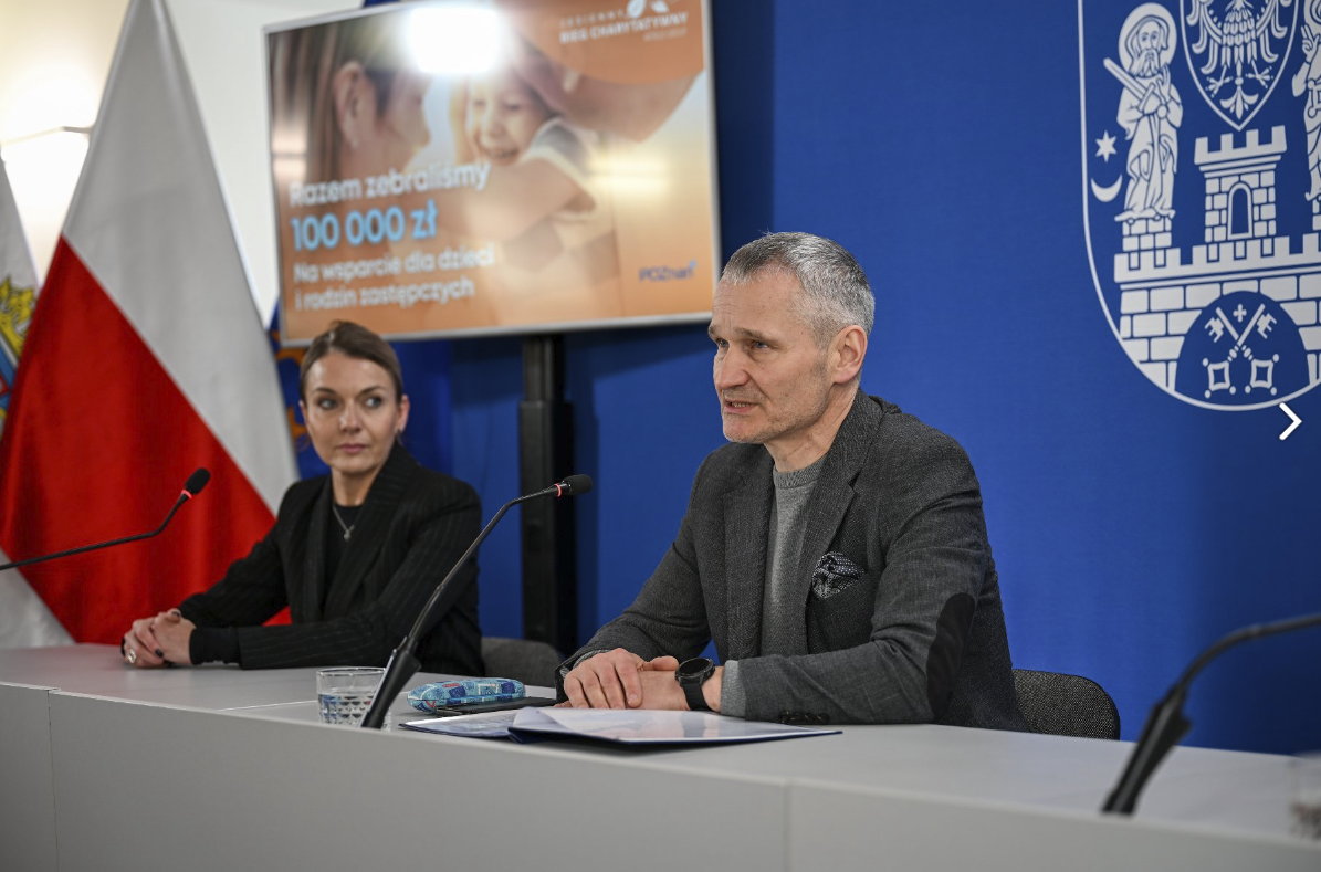 mężczyzna i kobieta przed mikrofonami, w tle niebieska ściana i biało-czerwona flaga