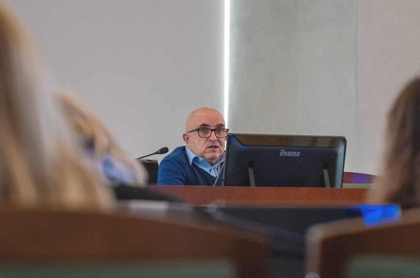 Mężczyzna za stołem prezydialnym przemawia w sali zwrócony do innych uczestników spotkania - więcej w treści artykułu - grafika artykułu
