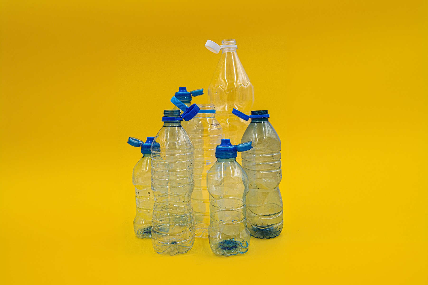 Na zdjęciu, na żółtym tle, widać siedem pustych plastikowych butelek z odkręconymi, ale przywtierdzonymi nakrętkami