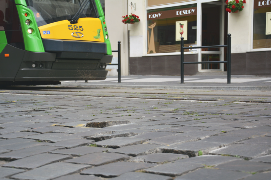 Na zdjęciu nawierzchia z nierówną kostką, w tle część tramwaju i wejście do kawiarni.