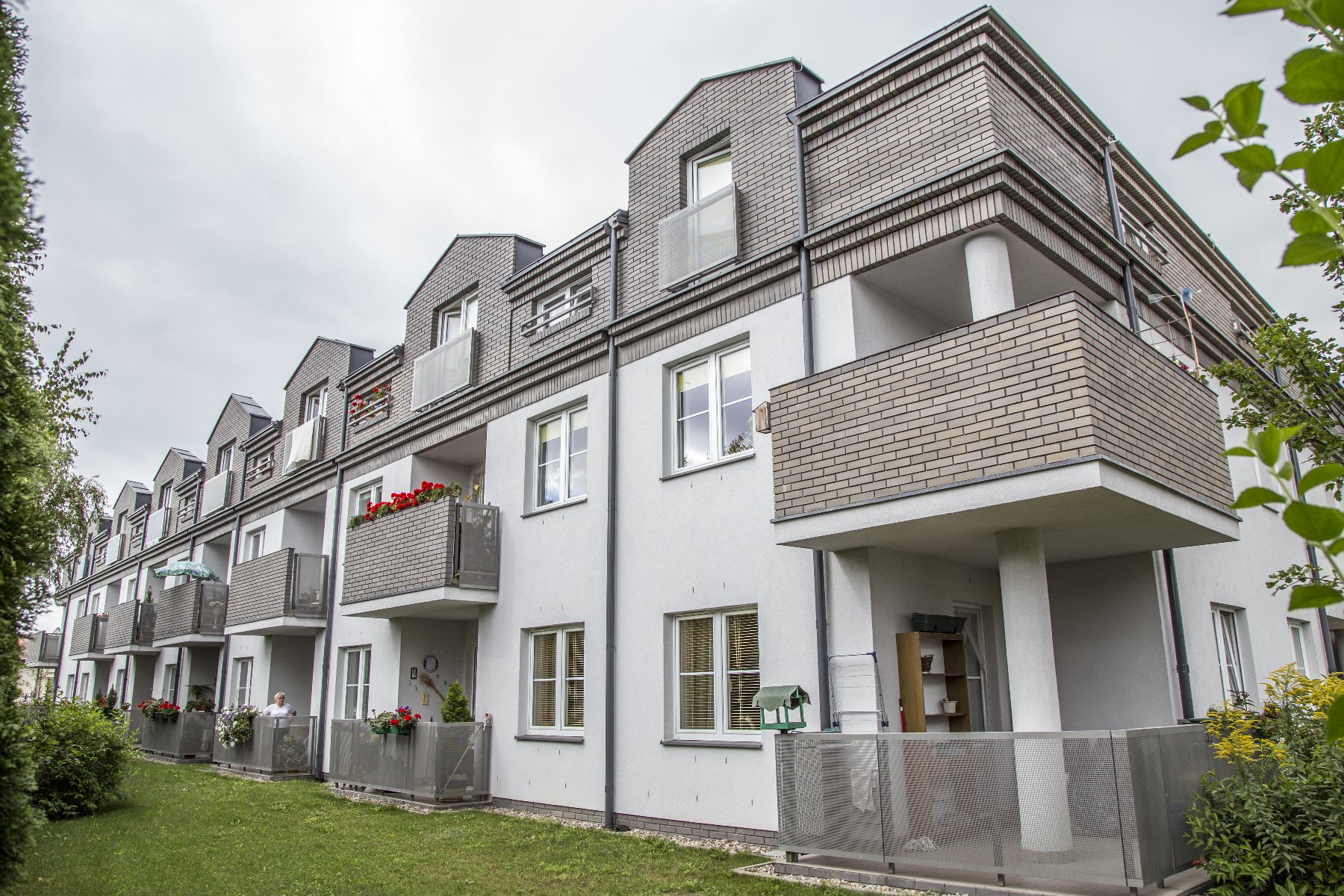 Na zdjęciu widać budynek, który ma 3 kondygnacje - parter i dwa piętra. Budynek widać od tyłu, nie od frontu. Przy każdym z mieszkań w tym budynku jest balkon. Przed budynkiem znajduje się trawnik.