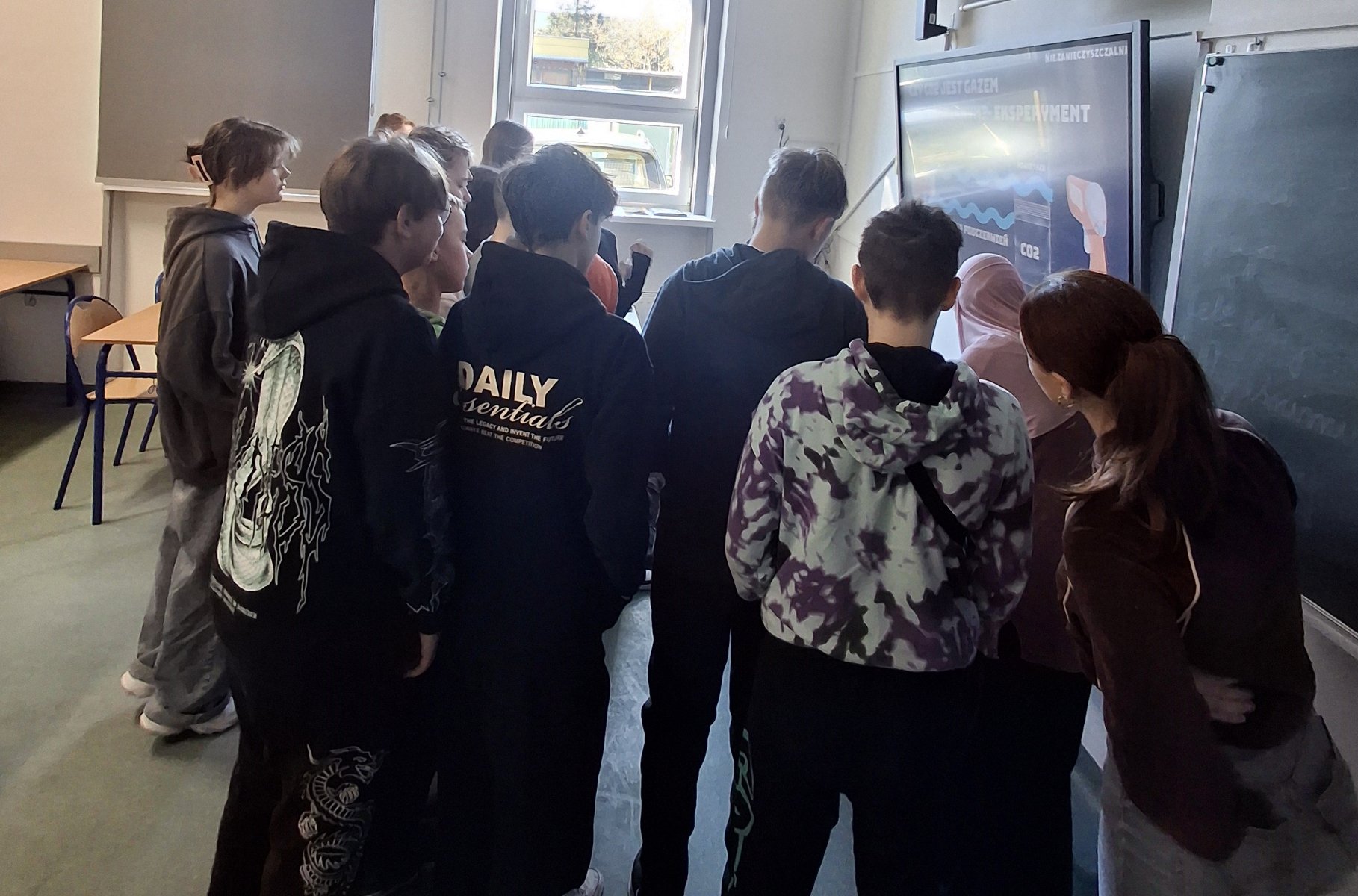 Na zdjęciu widać grupę młodzieży stojącą przy szkolnej tablicy i obserwującą działania niewidoczne w kadrze