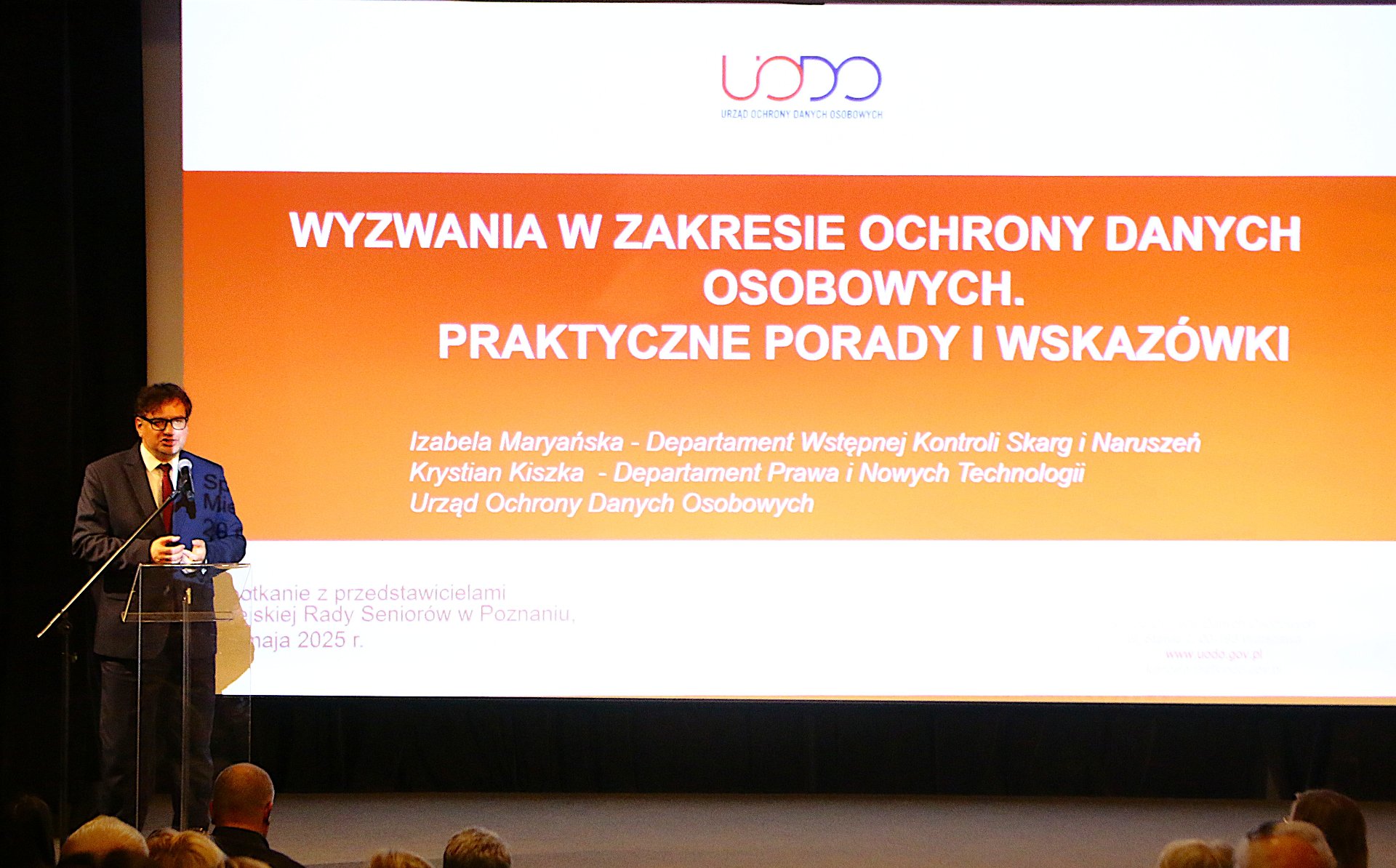 Na zdjęciu widzimy Pana prelegenta na tle slajdu z prezentacji omawianej na spotkaniu. Slajd w kolorze pomarańczowym.