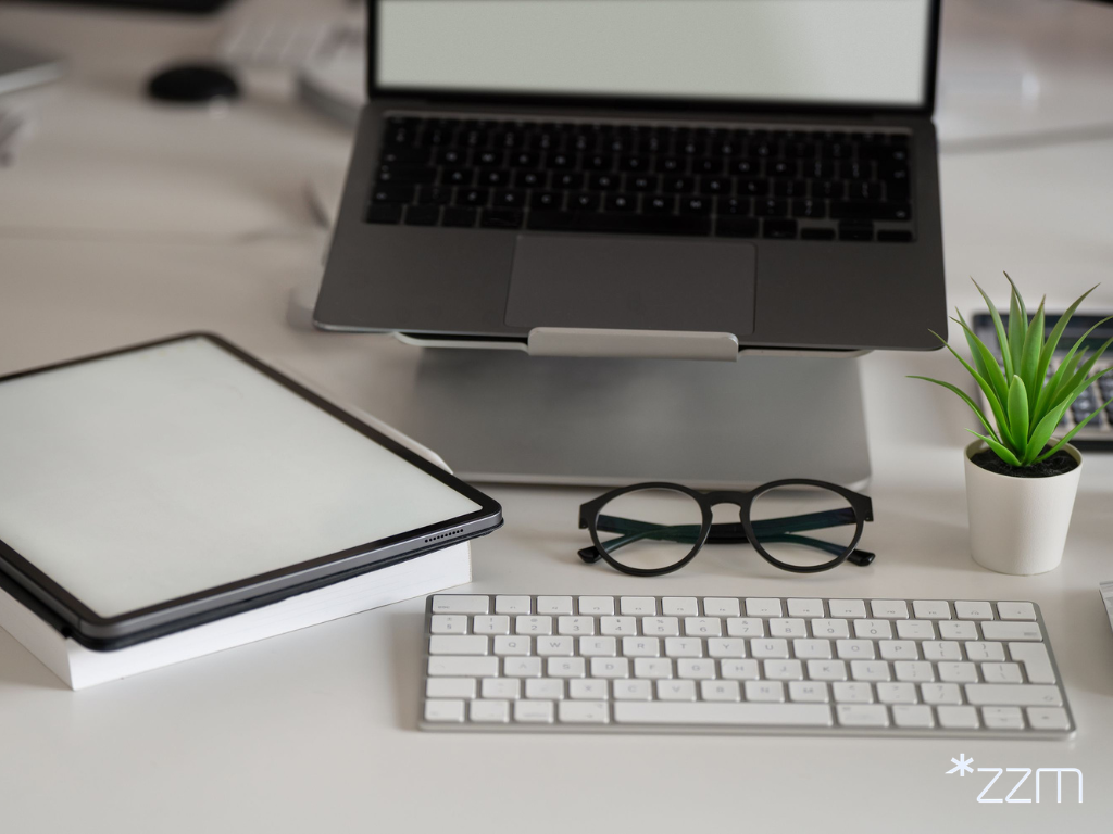 Nowoczesne stanowisko pracy na białym biurku. Na podwyższeniu stoi otwarty laptop z jasnym, pustym ekranem, a przed nim leżą biała klawiatura i czarne okulary. Po lewej stronie znajduje się tablet oparty na książce, a po prawej niewielka zielona roślina w białej doniczce oraz szklanka z wodą.